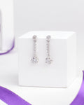 The Barely Bling Silver Earrings by Chandrani Pearls feature round and teardrop-shaped clear CZ stones for easy-care luxury, shown on a white cube with a purple ribbon beside them.