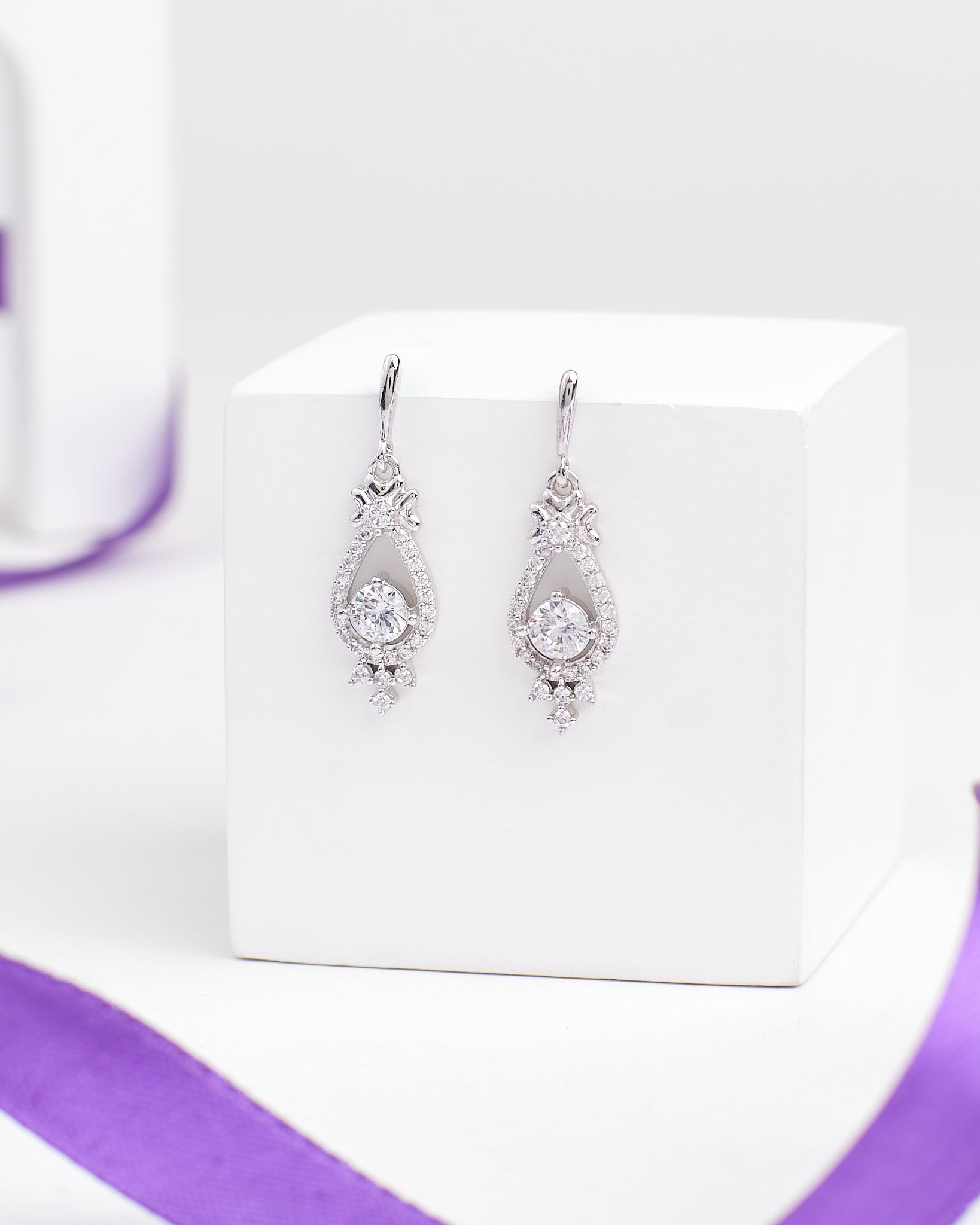 The Linger Longer CZ Silver Earrings by Chandrani Pearls feature teardrop-shaped luminous silver drops with clear gemstones, displayed on a white block and accented by a nearby purple ribbon.