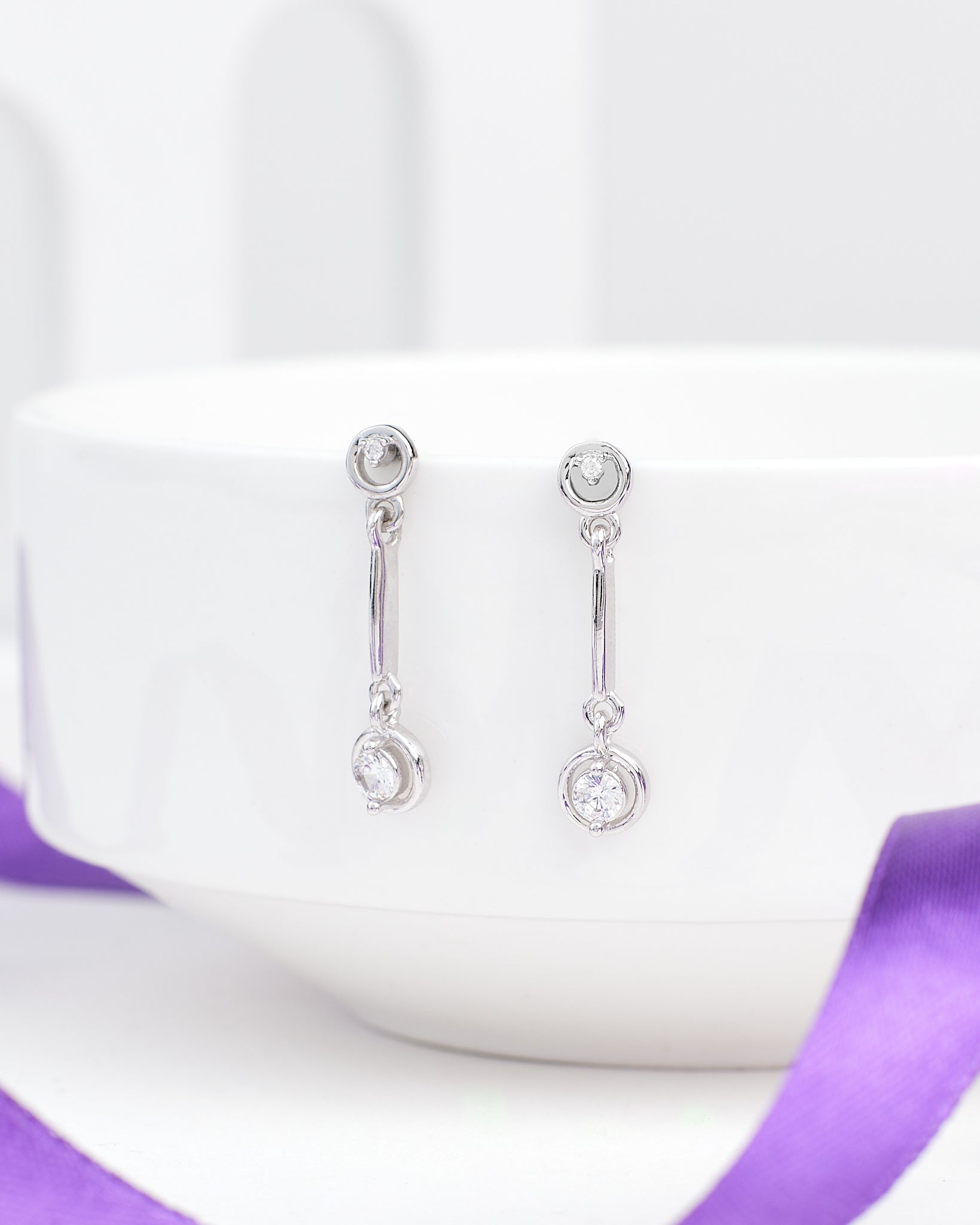 The Low Key Luxe CZ Silver Earrings from Chandrani Pearls feature round clear stones and softly shine, shown on a white ceramic bowl with a purple ribbon—perfect for adding relaxed style to any look.
