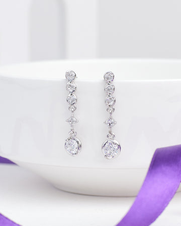 Date Night Lite Silver Earrings by Chandrani Pearls feature multiple round and teardrop-shaped clear gemstones set in silver. Styled against a white bowl with a purple ribbon, they're perfect to add sparkle to your next evening out.