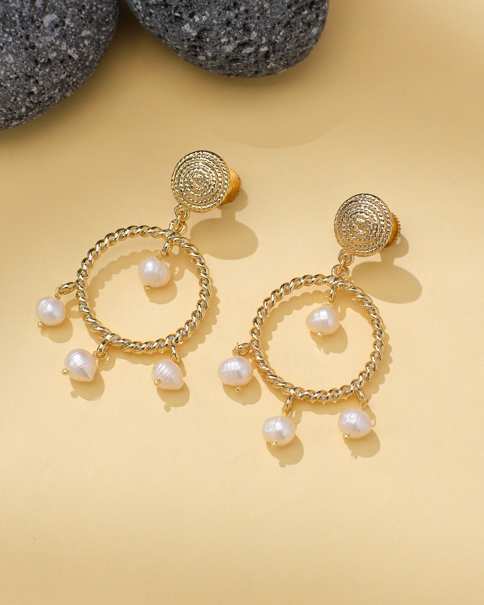 Halo Teardrop Earrings with Pearl Center by Chandrani Pearls are shown on a light yellow surface beside textured gray stones. Each gold earring features a teardrop shape with a pearl at the center.