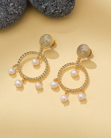 Halo Teardrop Earrings with Pearl Center by Chandrani Pearls are shown on a light yellow surface beside textured gray stones. Each gold earring features a teardrop shape with a pearl at the center.