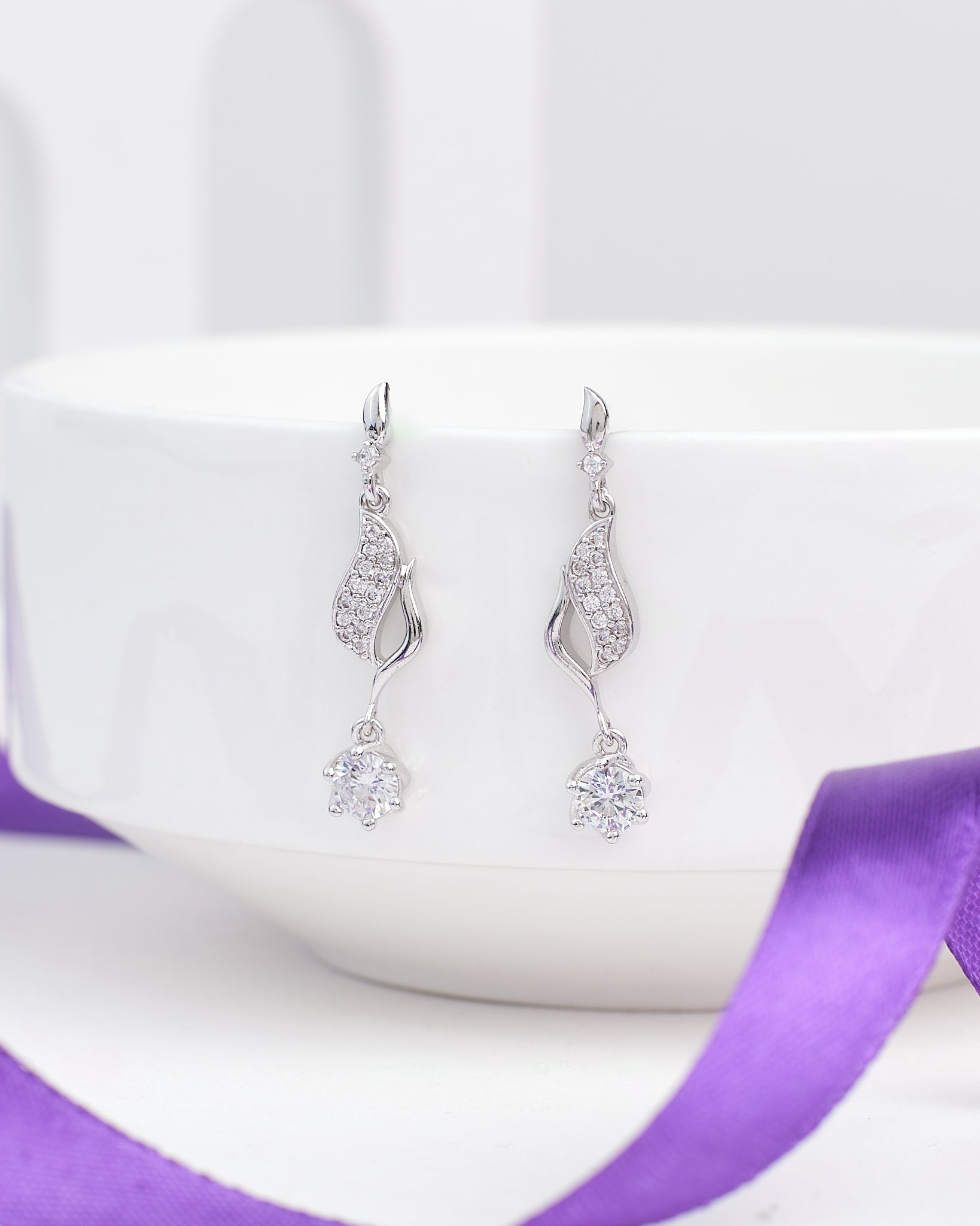 The Statement CZ Earrings in Polished Demifine Silver by Chandrani Pearls hang over a white bowl, sparkling with CZ stones, with a purple ribbon in the foreground.