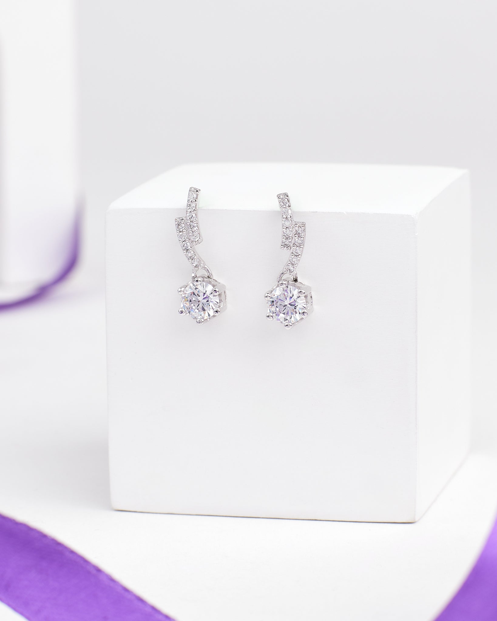 The Romantic CZ Drop Earrings in Demifine Silver by Chandrani Pearls gleam with sparkling stones, elegantly presented on a white stand with a purple ribbon backdrop.