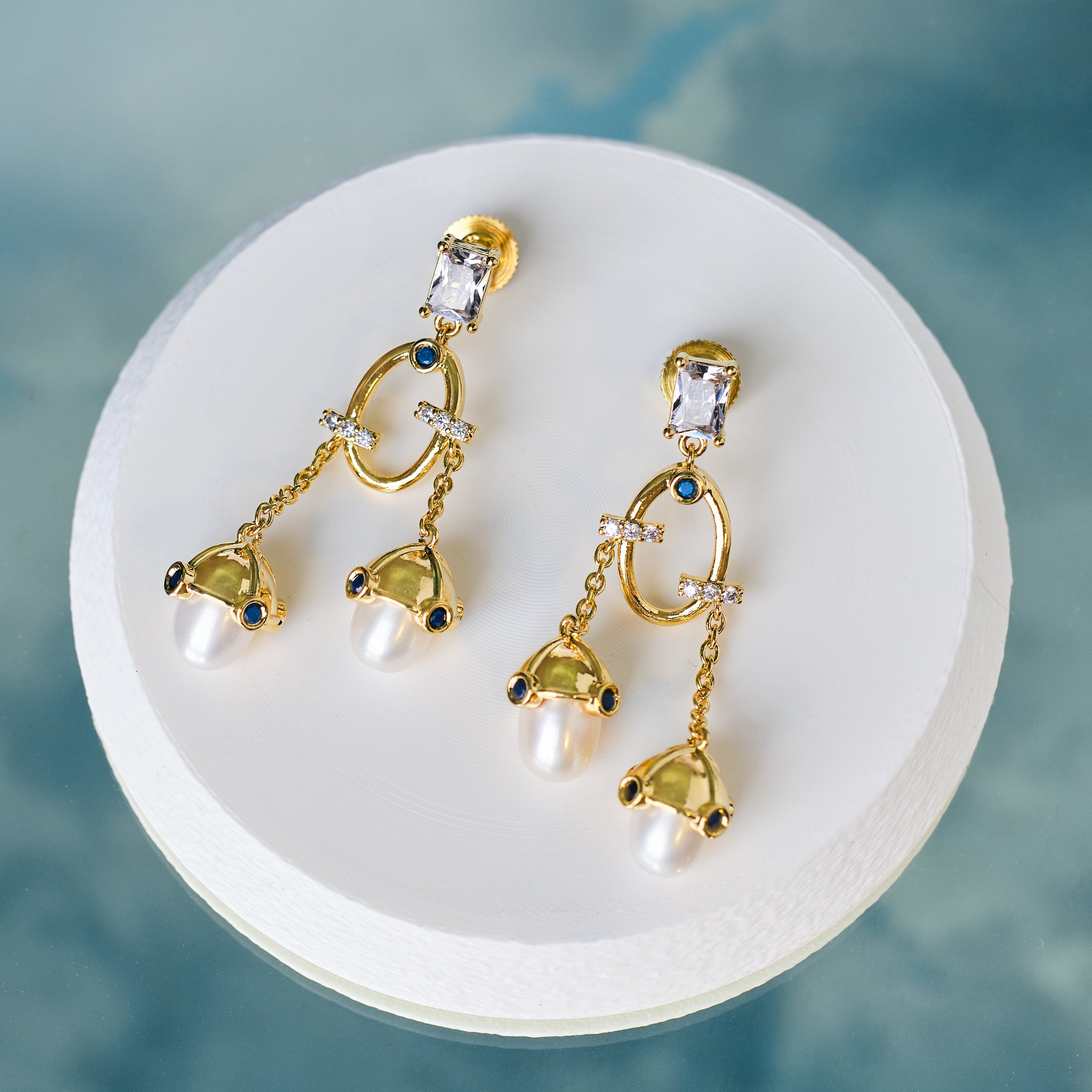 Chandrani Pearls' Stylish Handcrafted Pearl Earrings with CZ Sparkle are displayed on a round white platform against a blue background—perfect for adding everyday elegance.