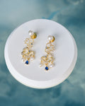 Gorgeous CZ & Pearl Stud Earrings Handcrafted by Chandrani Pearls feature decorative loops with pearl accents, clear crystals, and blue teardrop gemstones, elegantly displayed on a round white platform.