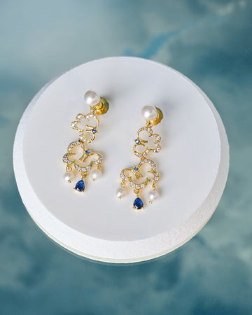Gorgeous CZ & Pearl Stud Earrings Handcrafted by Chandrani Pearls feature decorative loops with pearl accents, clear crystals, and blue teardrop gemstones, elegantly displayed on a round white platform.