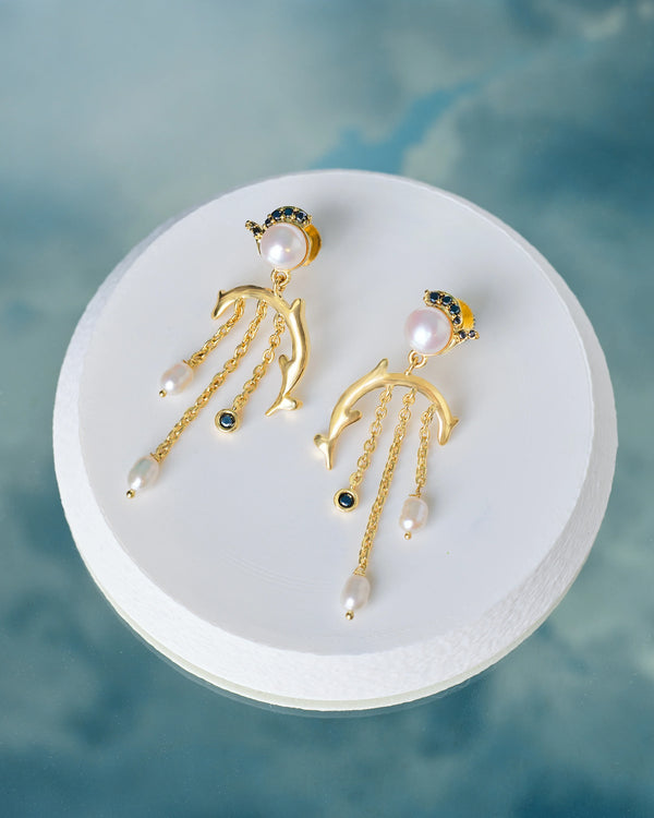 The Handcrafted Pearl & CZ Stone Earrings Elegant by Chandrani Pearls feature dolphin-shaped details, pearl accents, and dangling chains, shown on a round white platform against a blue backdrop.