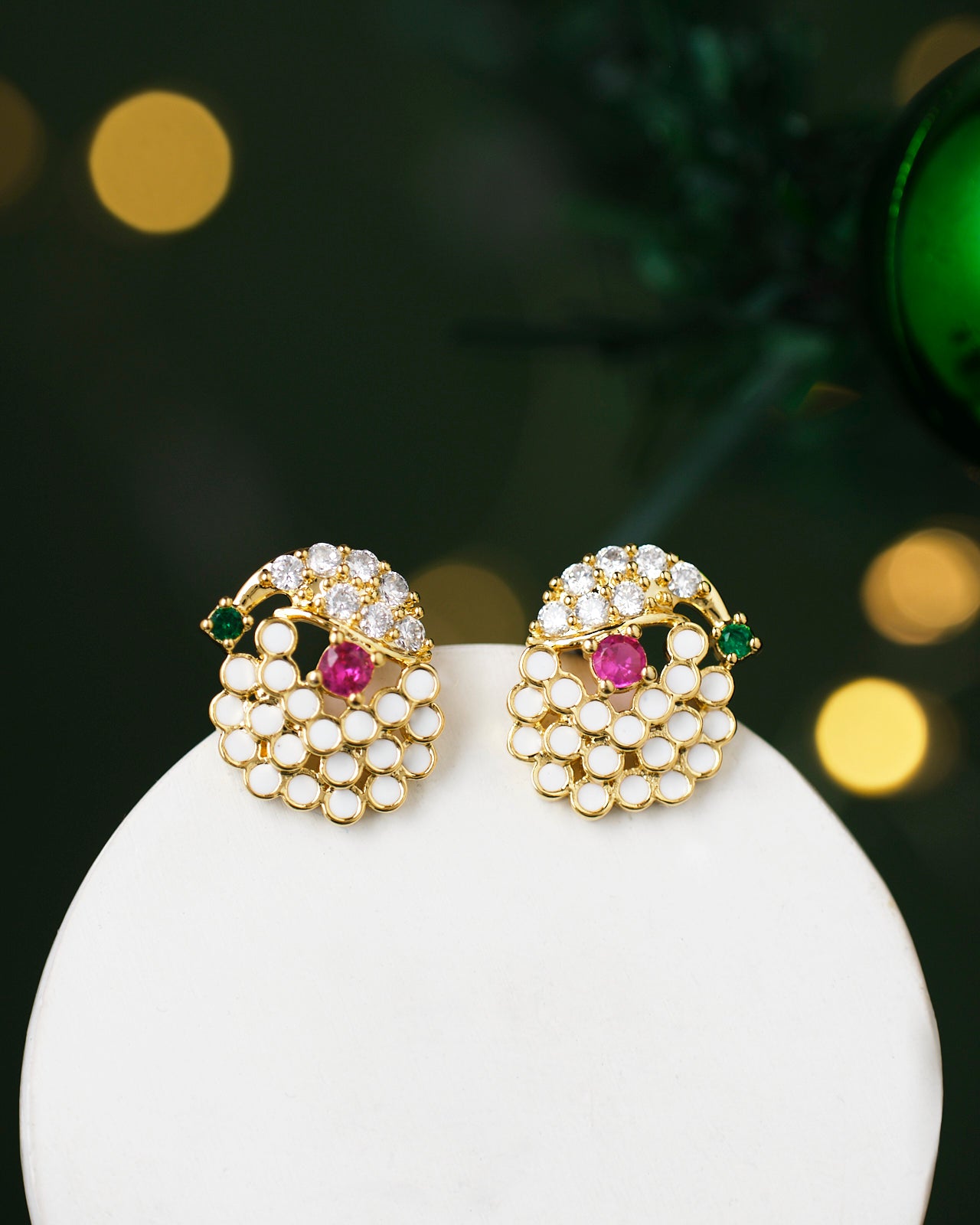 The Anurita Multi Stone Earrings by Chandrani Pearls feature gold earrings with pearl-like clusters, pink and green gemstones, displayed on a white stand against a backdrop of blurred lights.