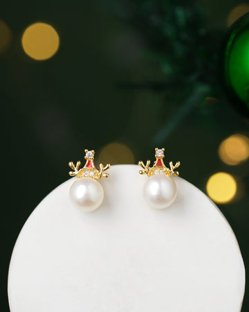 Blushing Dandelion Pearl Earrings by Chandrani Pearls feature gold reindeer antler designs with pearls and red accents, set against a blurred dark green background with glowing lights.